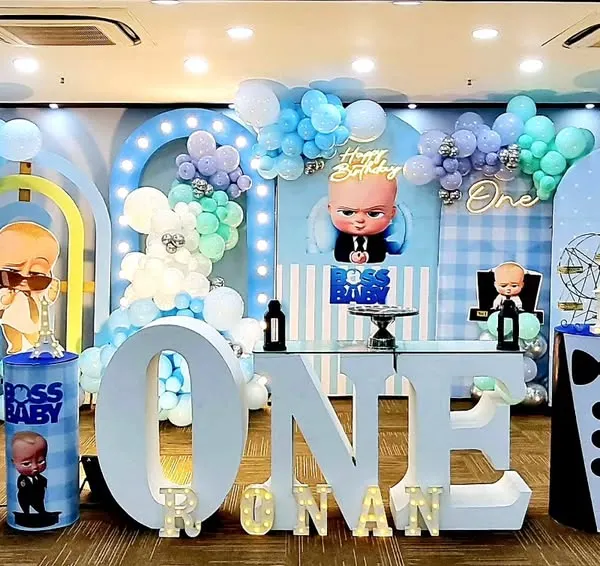 A spectacular Boss Baby themed first birthday setup, featuring large marquee letters for 'ONE', custom character cutouts, and an elaborate balloon arrangement in shades of blue, mint, and silver. This shows a complete themed stage design perfect for milestone celebrations.