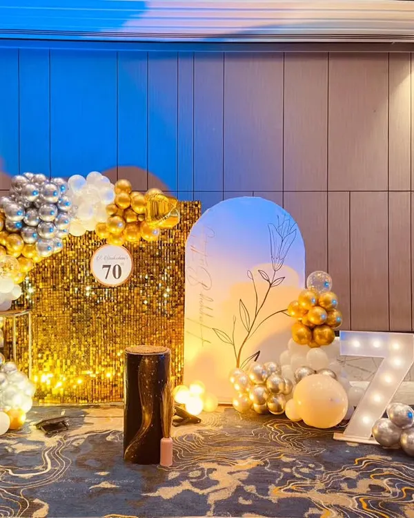 A glamorous 70th birthday celebration featuring a gold sequin shimmer wall, a silver and gold balloon arch, and large light-up marquee numbers.