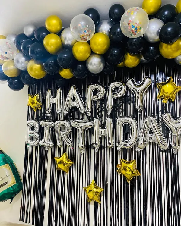 A classic and festive "Happy Birthday" setup perfect for any home party. This decoration features a black fringe curtain, a silver foil "Happy Birthday" banner, and a ceiling garland of black, gold, and silver balloons.