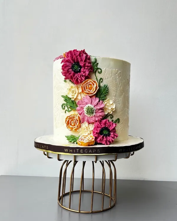 This tall celebration cake features intricate, hand-piped buttercream flowers in vibrant pinks and oranges, set against a subtly textured white frosting. It's a perfect example of the elegant, handcrafted wedding and anniversary centerpieces I love to create.