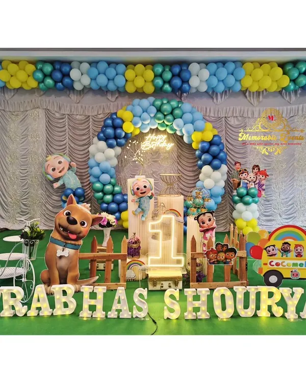 A grand Cocomelon theme for a first birthday party, complete with character cutouts, a custom backdrop, and a balloon arch in the show's signature colors. The setup includes a light-up number '1' and the birthday boy's name in marquee letters.