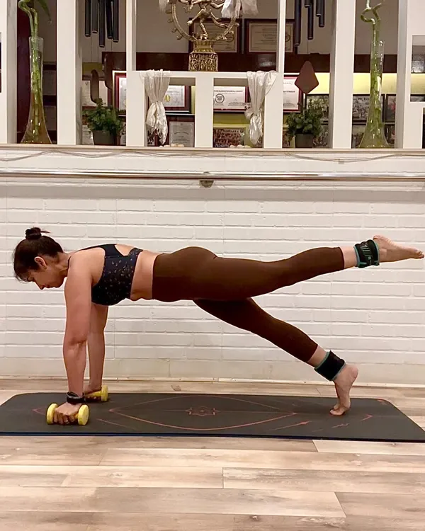 Pilates is about precision and control. Here, I am holding a plank with a leg lift, using ankle weights for added resistance and light dumbbells to engage my arms and shoulders. This single pose is a full-body exercise that strengthens the core, glutes, and upper body simultaneously.