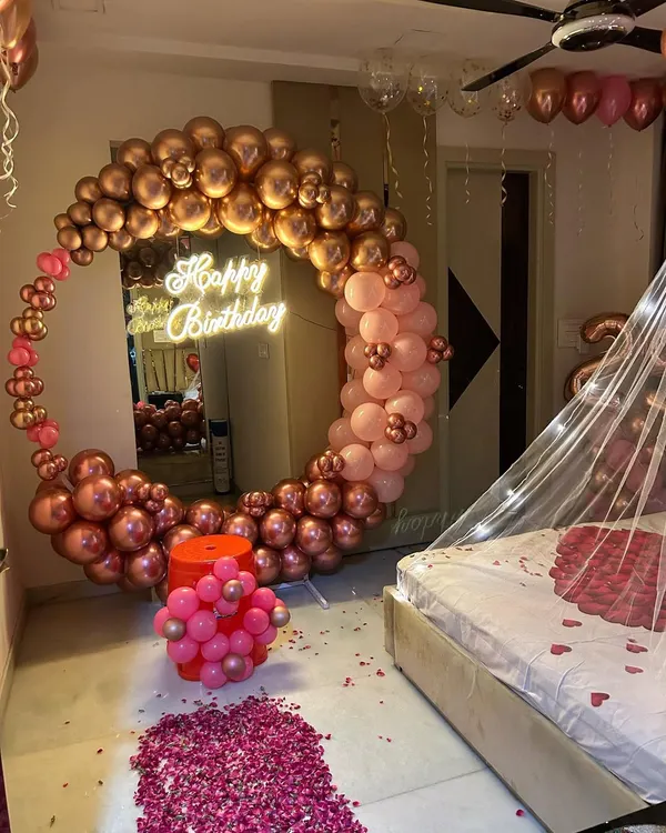 A romantic birthday room decoration featuring a large balloon ring in chrome rose gold. The setup includes a 'Happy Birthday' neon sign and a path of rose petals, creating a perfect surprise.