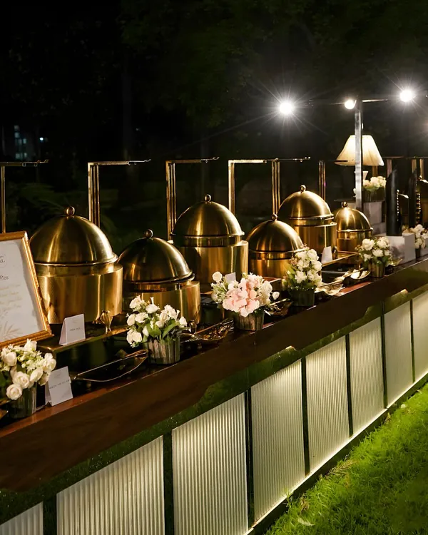 An elegant outdoor buffet line at night. The station is illuminated with warm lighting, highlighting the row of gleaming brass chafing dishes and creating a sophisticated and inviting atmosphere on the lawn.