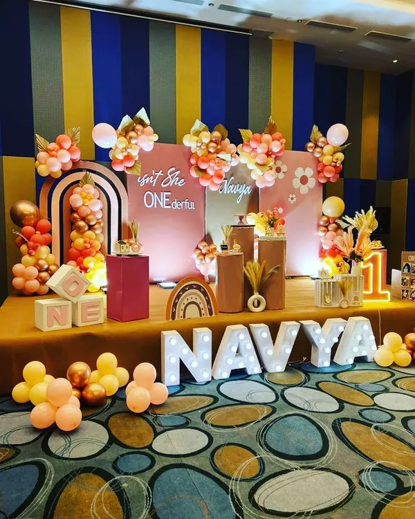 A "Boho Rainbow" themed first birthday party for Navya. This stage setup features pastel balloon arrangements, a custom backdrop, and large marquee lights for the number '1' and the name.