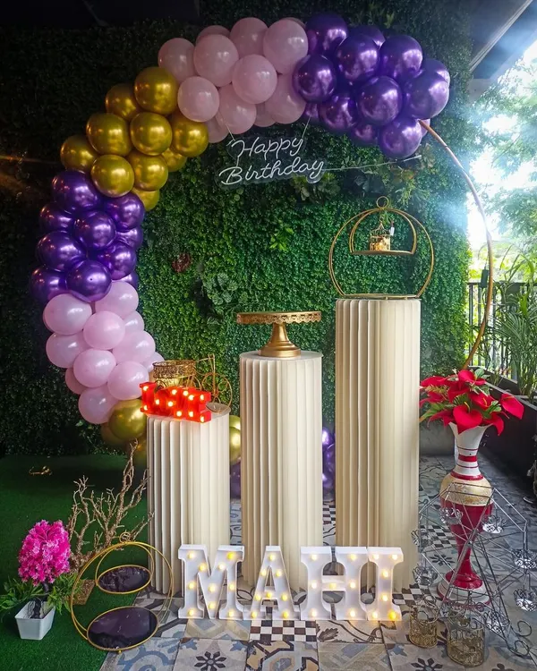 We combined natural and modern elements for this birthday setup. A lush greenery wall serves as the perfect base for a colorful balloon arch in purple, pink, and gold, complete with a neon sign and elegant pedestals for the cake.