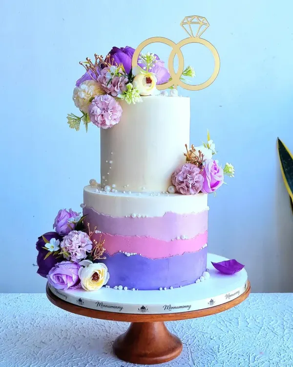 A two-tiered wedding or engagement cake with a purple ombre effect. This design is decorated with artificial flowers and a gold ring topper, but I teach how to create similar flowers from sugar.