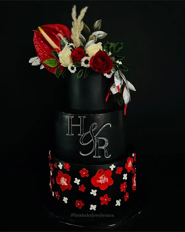 A black wedding cake is a statement. This dramatic three-tiered creation features a cascade of red and white sugar flowers and a custom monogram, proving that wedding cakes can be both bold and beautiful.