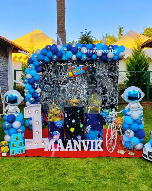 An exciting outdoor space-themed first birthday party. The setup features a large sequin backdrop, balloon astronaut figures, a custom milestone board, and even a miniature car for the birthday boy.