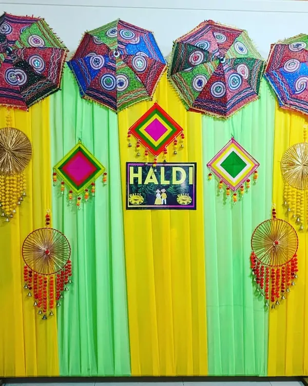 A colorful and traditional Haldi backdrop. This setup features yellow and green drapes, decorative umbrellas, and handcrafted hangings for a festive look.