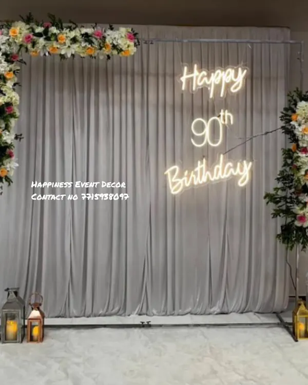 An elegant and classy setup for a 90th birthday celebration. This decor features a simple frame with a white curtain backdrop, floral arrangements, lanterns, and a custom neon sign.
