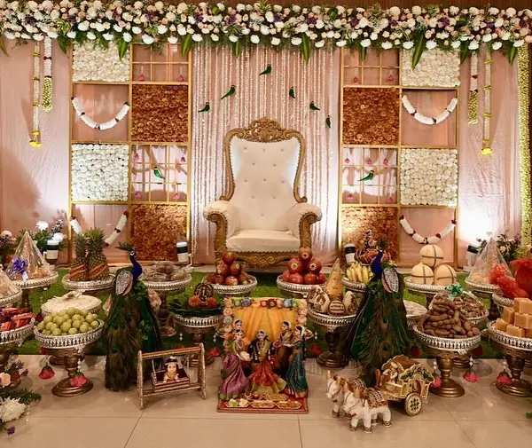 A complete traditional setup for a special occasion. We designed this elegant stage with a floral grid backdrop, a regal throne, and our signature 21-plate offering display featuring fruits, sweets, and decorative peacocks.