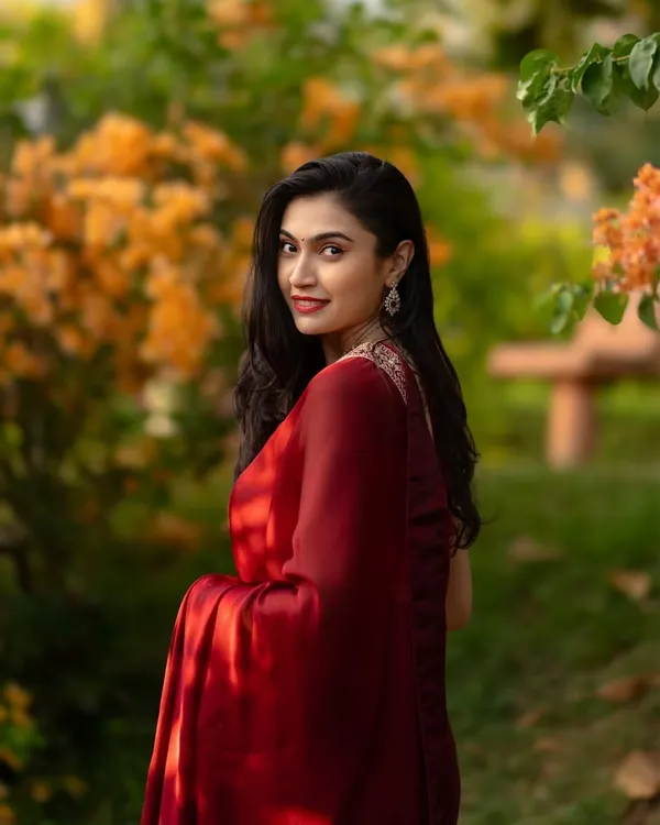 An elegant portrait of a woman in a deep red saree, looking over her shoulder with a gentle smile against a backdrop of blooming orange flowers.