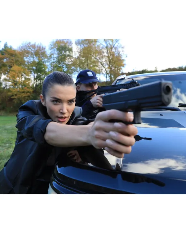 A dynamic action shot of Amy Jackson as Patricia Novak, taking cover behind a vehicle. The sleek black leather jacket and pulled-back hair create a practical and intimidating look for her character.