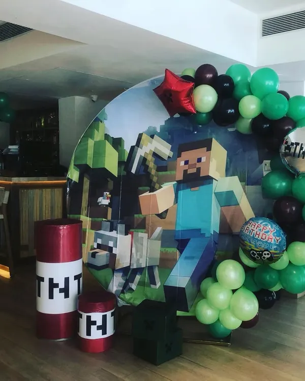 A full Minecraft-themed party setup. This includes a large printed backdrop with game characters, themed props like TNT barrels, and a balloon garland in signature green and black colors.