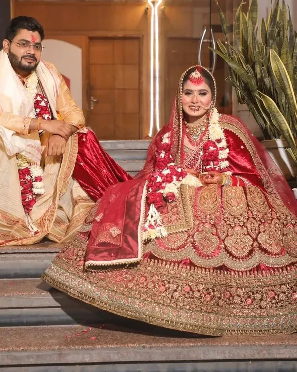 A regal portrait of the bride and groom on their wedding day. The bride is seated on the steps, allowing her magnificent red and gold lehenga to be displayed in its entirety, while the groom sits beside her in his traditional attire. This posed shot captures the grandeur of the occasion.