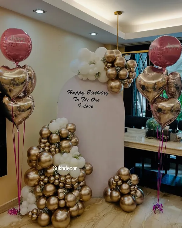 An elegant birthday backdrop featuring a large arch panel with the message 'Happy Birthday To The One I Love'. The setup is decorated with beautiful chrome gold and white balloon arrangements and heart-shaped foil balloons.