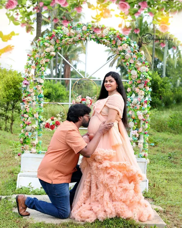 A beautiful couple's portrait under a vibrant floral arch. The partner's gentle kiss on the baby bump is a classic pose that captures the love and excitement of this special time.