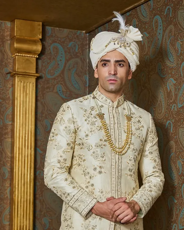 An ivory and gold sherwani is the epitome of classic groomswear. This piece features delicate, all-over floral embroidery, paired with a matching safa and a traditional gold necklace for a truly royal look.