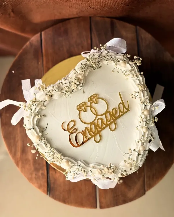 An elegant heart-shaped engagement cake. It's adorned with delicate baby's breath, white ribbons, and a golden "Engaged" topper for a classic, romantic look.