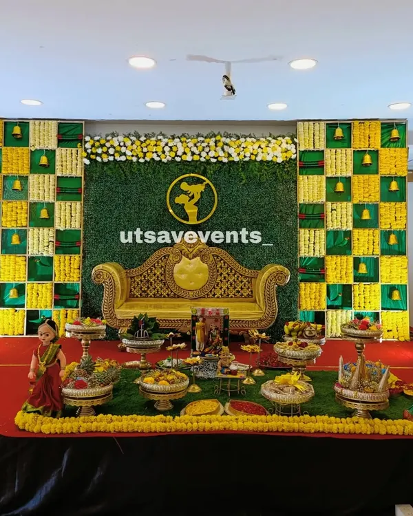 This is a complete baby shower setup, showing how all the elements come together. I've used a traditional checkerboard floral backdrop in auspicious yellow and green, an ornate golden sofa for the mother-to-be, and a full display of 21 ceremonial plates on a green turf stage.