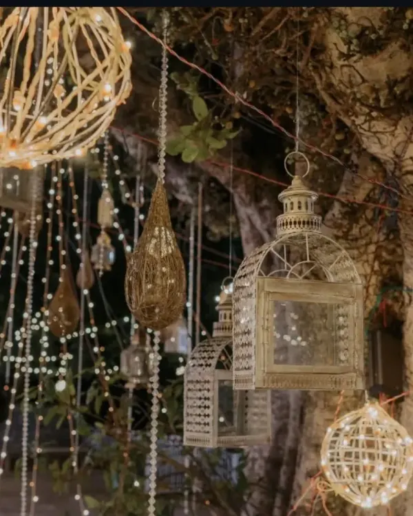 We create magical ambiance with creative lighting. Here, a collection of hanging lanterns, including birdcages and woven orbs filled with fairy lights, illuminates an outdoor wedding venue.