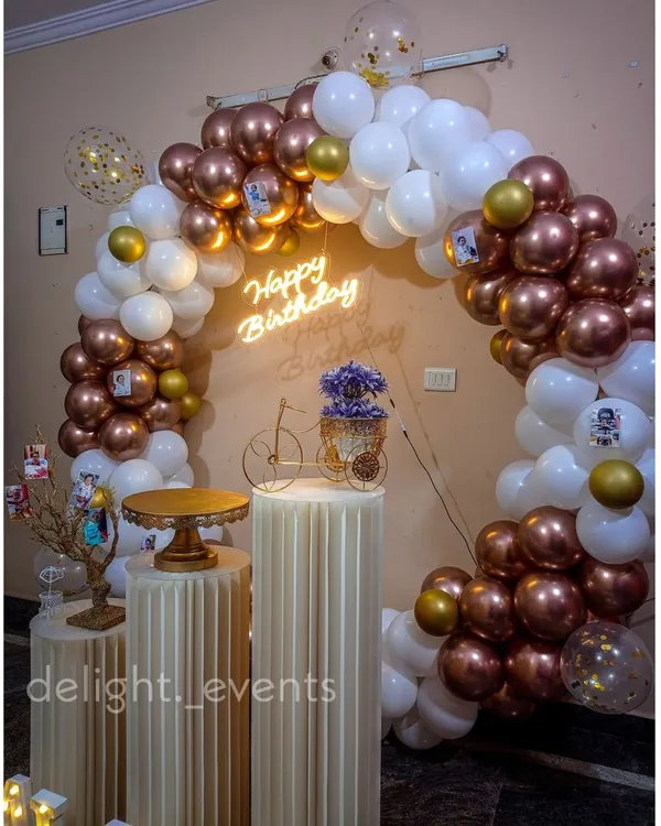 An elegant and popular choice, this full circular balloon arch combines metallic rose gold and classic white balloons. It's paired with a neon 'Happy Birthday' sign and fluted pedestals, creating a chic and modern photo corner.