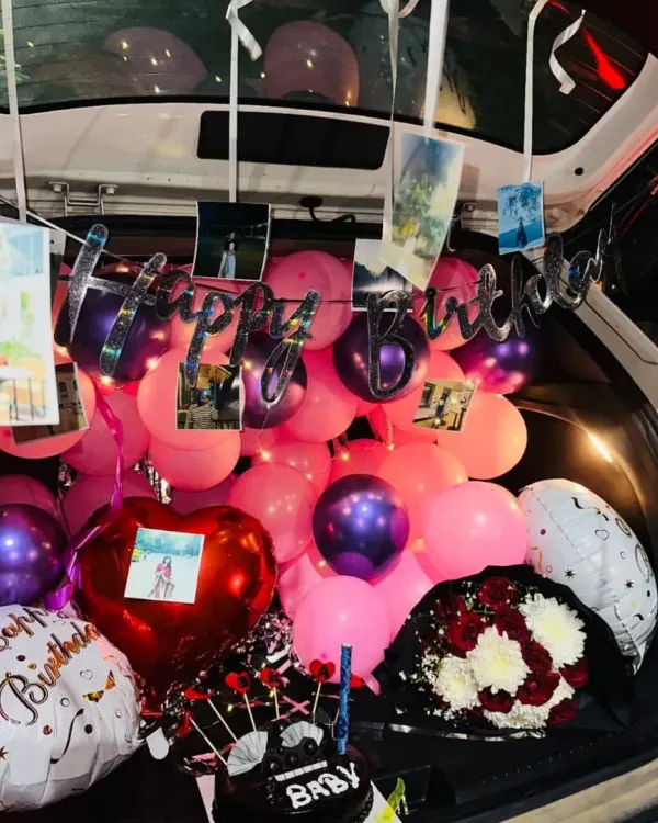 A car trunk filled to the brim for a birthday surprise, including pink and purple balloons, a 'Happy Birthday' banner, hanging photos, a cake, and a bouquet.