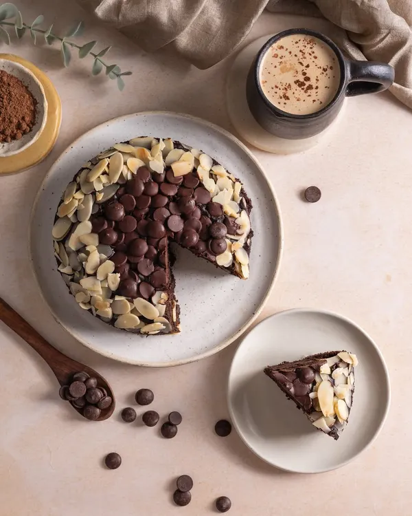 A top-down shot of a rich chocolate almond cake, with a slice cut out to show the texture inside. This flatlay style is perfect for menus and social media, showing the cake, a coffee, and the ingredients.