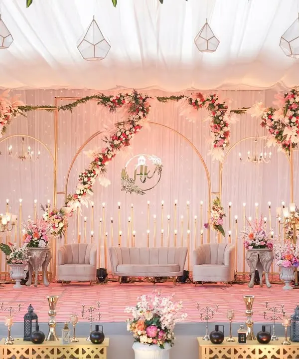 A truly regal reception stage in blush pink tones. The setup features elegant seating, cascading floral arrangements, and delicate candle lighting, all framed by custom golden arches for a majestic feel.