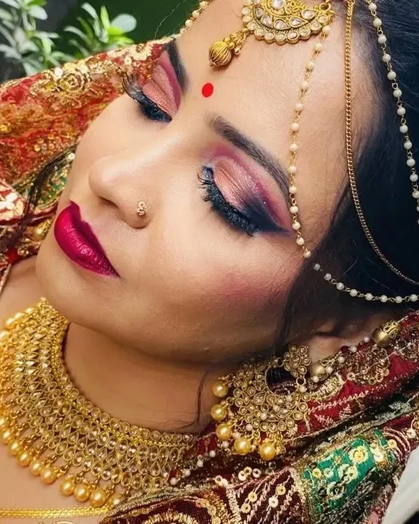 This North Indian bridal makeover features a dramatic smokey eye with pink and gold tones, paired with a bold magenta lip. The complete look, including the intricate matha patti and jewelry, creates a radiant and unforgettable bridal appearance.