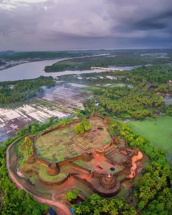 An aerial panorama of Mirjan Fort near Kumta, captured under a dramatic monsoon sky. The drone's perspective reveals the fort's strategic location, nestled amidst lush greenery and the winding Aghanashini river.