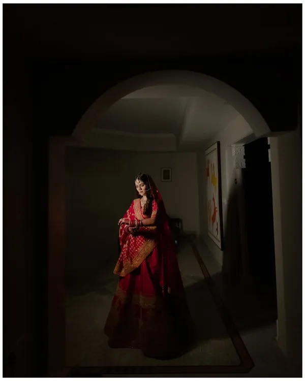 A dramatic bridal portrait using shadow and light. The bride, in her radiant red lehenga, is framed by an archway, creating a sense of mystery and timeless beauty.
