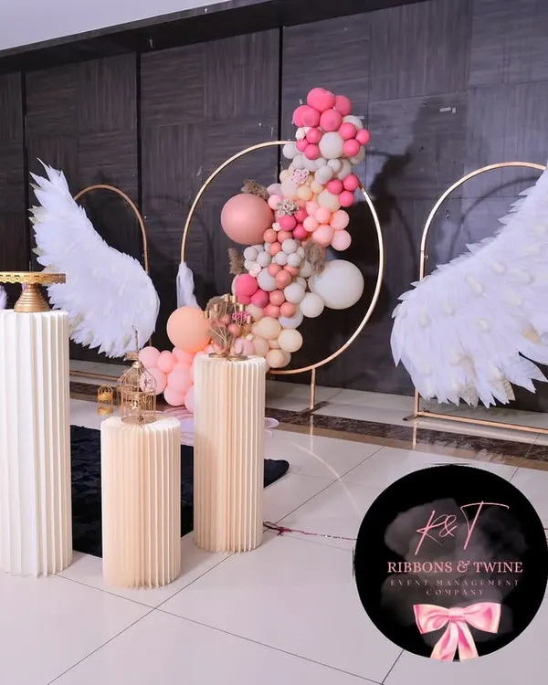 A beautiful baptism setup featuring angel wings, a central gold ring with a balloon garland, and fluted plinths for the cake and other displays. The color scheme of pink, peach, and white creates a soft and celebratory feel.