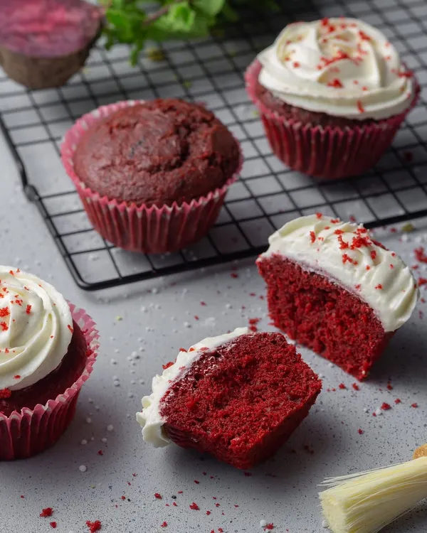 Red velvet cupcakes with cream cheese frosting. The composition includes a cut-open cupcake to show the moist, vibrant crumb, a key detail for bakery photography.
