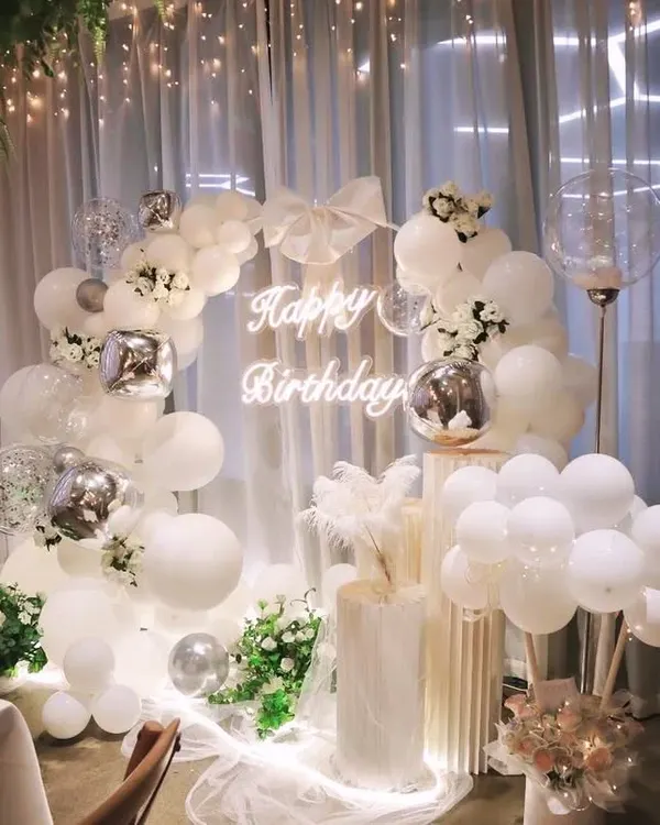 An ethereal and dreamy all-white birthday theme. This setup features a beautiful white balloon arch, a sheer curtain backdrop, a neon sign, and delicate floral accents for a truly elegant and sophisticated celebration.