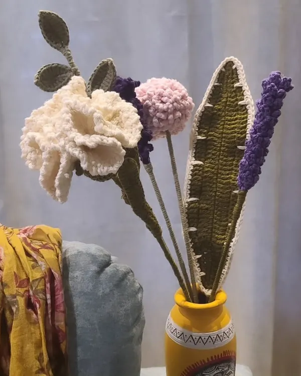 A beautiful, long-lasting crochet flower arrangement in a bright yellow vase. This handmade bouquet includes a detailed white carnation, a fluffy pink pom-pom, a sprig of purple lavender, and textured green leaves, offering a cheerful and permanent decorative accent.