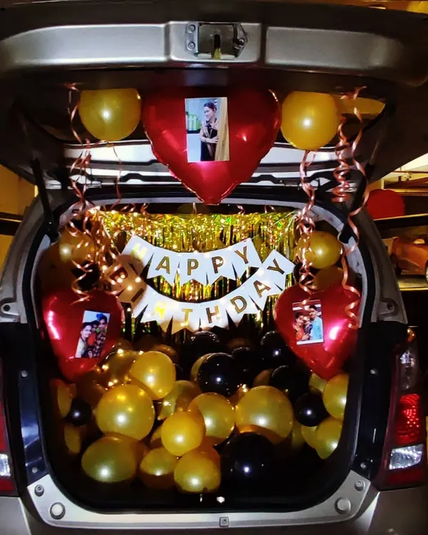 A personalized car trunk birthday surprise. This setup includes black and gold balloons, a 'Happy Birthday' banner, and custom heart-shaped balloons featuring photos of the birthday person.