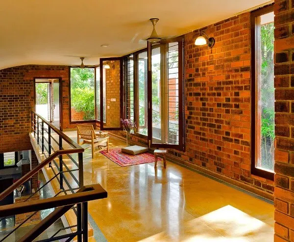 The upper-level corridor at the Vidyut Kapoor Residence features a continuous exposed brick wall, complemented by large windows that offer views of the surrounding greenery. The polished yellow oxide flooring reflects the warm tones and natural light.