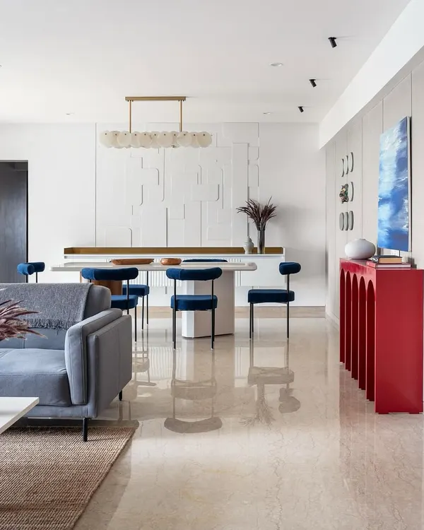 The living and dining area of 'Vishu Home' showcases a bold design statement. A striking red arched console table adds a vibrant pop of color against the neutral textured wall, while the blue dining chairs provide a cool contrast.
