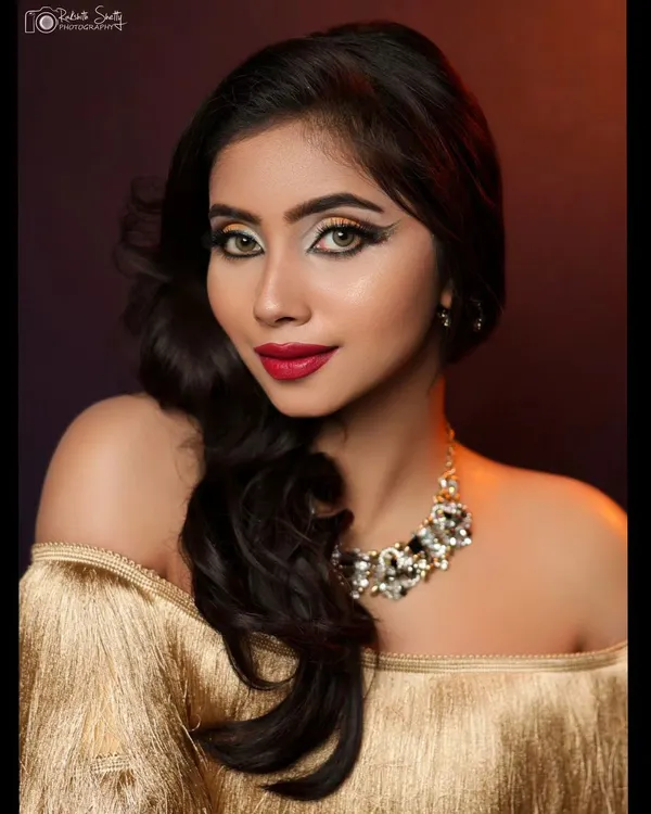 A glamorous studio portrait with dramatic lighting. The focus is on the model's striking makeup, statement necklace, and the texture of the gold off-shoulder top.