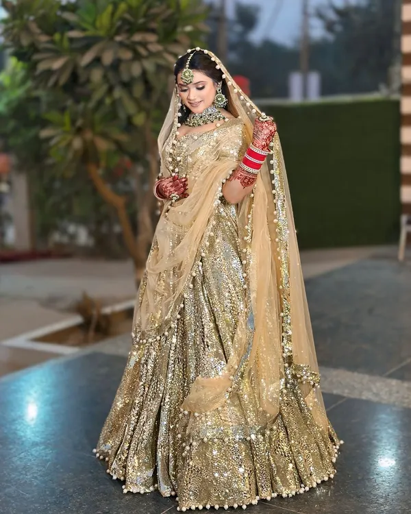 A full-length view of a magnificent golden sequin lehenga. The pearl-bordered dupatta and green-accented jewellery add a touch of classic elegance.