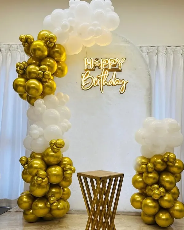 An elegant birthday decoration in gold and white. This setup features an organic balloon arch, a "Happy Birthday" neon sign against a white panel, and a stylish pedestal for the cake.