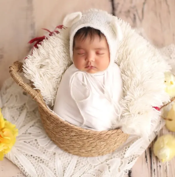 A vision in white. This classic newborn portrait features a baby sleeping peacefully in a basket, wearing an adorable bear bonnet and surrounded by soft, spring-like details.