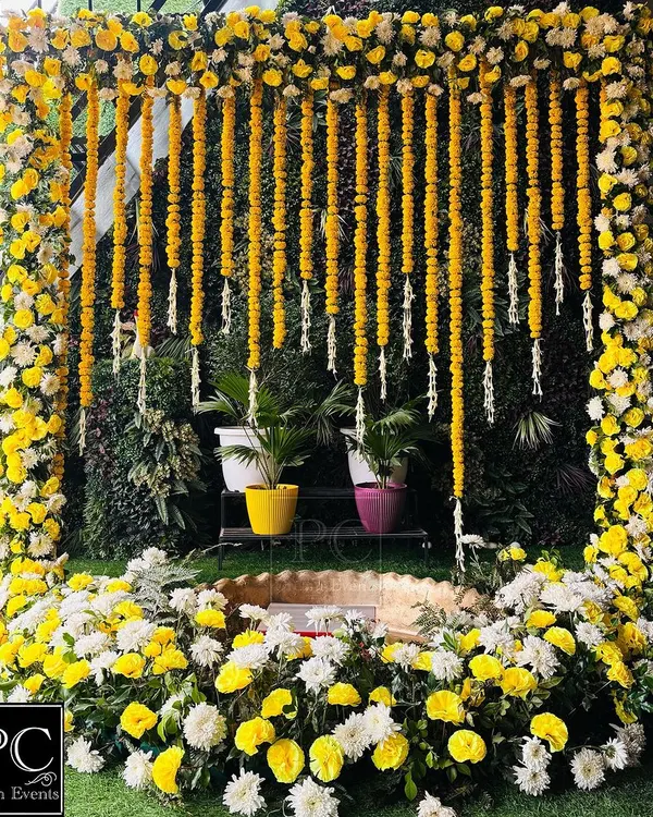 A classic Haldi setup featuring a frame of yellow and white flowers with hanging marigold garlands. The central seating area is designed for the ceremony, surrounded by lush greenery for a fresh, natural feel.