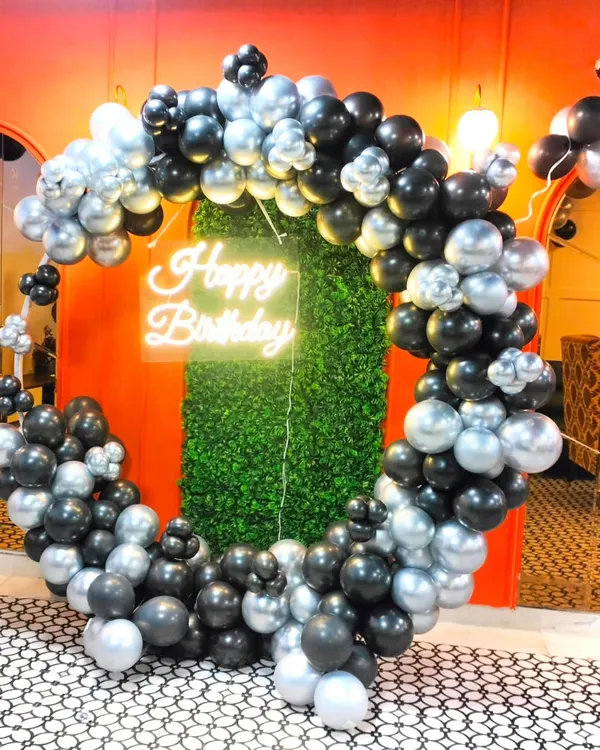 This is a great example of a sophisticated party backdrop for adults. A full ring of black and silver chrome balloons frames a green foliage wall, with a neon "Happy Birthday" sign in the center, creating a perfect spot for selfies and group photos.