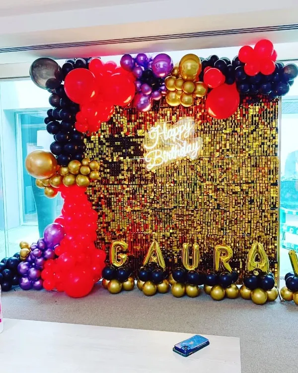 A vibrant and glamorous birthday backdrop. This setup features a gold shimmer wall framed by a dramatic balloon garland in red, black, purple, and gold, with a 'Happy Birthday' neon sign.