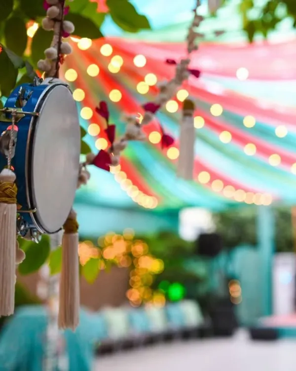 A beautiful detail from a festive decor setup, featuring a traditional dafli (tambourine) with tassels hanging from a tree. The background shows colorful drapes and string lights, creating a vibrant ambiance.
