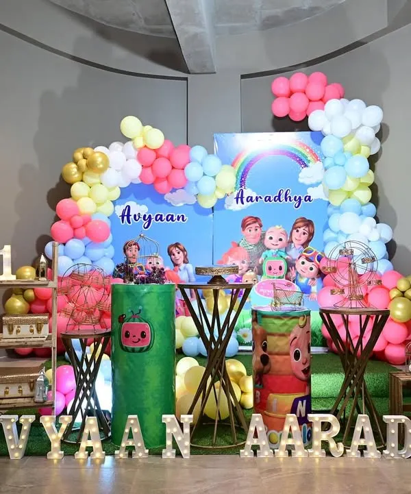 A bright and cheerful Cocomelon theme for a joint birthday party. The setup includes a custom backdrop, character cutouts, and themed cake tables.