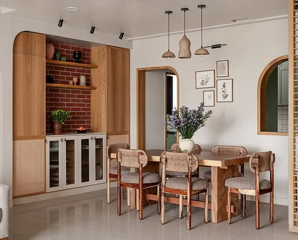 This dining area flows seamlessly from the living room, anchored by a custom live-edge dining table and rounded chairs. The crockery unit features a mix of fluted and cane details, while a brick-clad niche adds rustic charm.